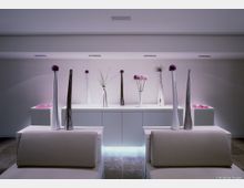 Minimalistischer Innenraum mit einer weiß beleuchteten Wand, darauf schmale Vasen in unterschiedlichen Größen mit dekorativen Pflanzen und lila Blüten. Im Vordergrund befinden sich schlichte cremefarbene Möbel, und der Boden besteht aus glänzenden Fliesen.