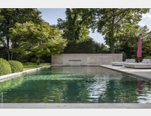 Moderner Außenpool in einem Garten mit gepflegter Rasenfläche und Bäumen im Hintergrund, umgeben von einer niedrigen Steinmauer. Auf der rechten Seite befinden sich Liegen und ein geschlossener violetter Sonnenschirm auf einer gepflasterten Terrasse.