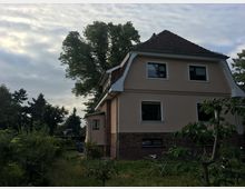 Ein zweistöckiges Wohnhaus mit hellbeigem Putz, einem dunklen Ziegeldach und teilweise verklinkerter Fassade, umgeben von einem Garten mit Bäumen und Sträuchern. Im Hintergrund sind weitere Bäume und ein bewölkter Himmel zu sehen.