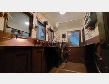 Führerstand einer historischen Lokomotive mit Steuerinstrumenten, Hebeln und mehreren Armaturen. Sichtbare Fenster und eine Tür geben den Blick auf den äußeren Bahnhofsbereich frei.