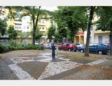 Ein kleiner, gepflasterter Platz mit geometrischem Muster, umgeben von Bäumen und Sträuchern. Im Hintergrund stehen mehrstöckige, gelb gestrichene Wohngebäude mit Balkonen, während parkende Autos und ein Brunnen den Vordergrund prägen.