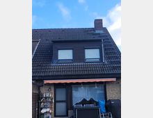 Außenansicht eines Reihenhauses mit einem dunklen Ziegeldach, zwei Dachfenstern mit Rollläden und einem gemauerten Schornstein. Im Erdgeschoss befinden sich eine Glastür, ein Fenster mit einer Gardine sowie eine Markise darüber, daneben stehen Gartenmöbel und Aufbewahrungsgegenstände.