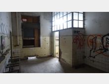 Ein verlassenes, heruntergekommenes Gebäude mit gefliesten Wänden in Gelb und Weiß, teils beschädigt und mit Graffiti versehen. Die Szenerie zeigt einen Raum mit großen, teilweise zerbrochenen Fenstern, einem offenen Durchgang und einem dreckigen Boden.