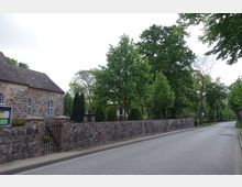 Eine Dorfstraße mit einer Steinmauer und einer kleinen Kirche aus Feldsteinen links im Bild. Die Umgebung ist von Bäumen und grüner Vegetation geprägt.