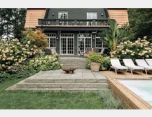 Gartenansicht eines Hauses mit dunkler Fassade und terrakottafarbenem Dach. Im Vordergrund befinden sich eine Steinterrasse mit Feuerstelle, Liegestühle auf einem Holzdeck neben einem Pool sowie üppige Blumenbeete mit Hortensien und großen Pflanzen.