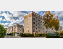 Mehrstöckige Wohngebäude mit hellen Fassaden und Balkonen, umgeben von grünen Hecken, Bäumen und einem Gehweg. Im Vordergrund befinden sich Müllcontainer und ein Halteverbotsschild.