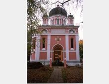 Rosa-weiß gestrichene orthodoxe Kirche mit markanten, dunklen Kuppeln, dekorativen Bögen und kleinen Türmchen; umgeben von einem Metallzaun und von Herbstbäumen gesäumt. Ein gepflasterter Weg führt zum Eingang mit einer verzierten Holztür.