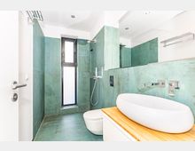 Modernes Badezimmer mit grünen Wand- und Bodenfliesen, einem rechteckigen Fenster mit Milchglas, einer offenen Regendusche und einem weißen, ovalen Waschbecken auf einer hellen Holzablage. Wandarmaturen und weitere minimalistische Ausstattungselemente ergänzen das Design.