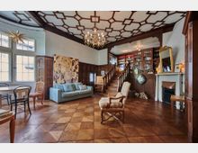 Wohnzimmer mit Holzboden und aufwendiger geometrischer Deckenverzierung, ausgestattet mit einer hellblauen Couch, einem gemusterten Sessel und einem Kamin. Eine Holztreppe führt zu einer erhöhten Bibliothek mit Bücherregalen, während große Fenster und Wandvertäfelungen das klassische Ambiente unterstreichen.