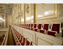 Innenansicht eines Konzertsaals mit einer Reihe roter Polstersitze auf einem oberen Rang. Die Wände sind mit Marmoroptik, goldenen Verzierungen und Büsten geschmückt; im Hintergrund eine glitzernde Kronleuchterbeleuchtung.