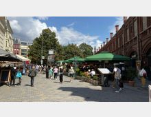 Straßenszene mit Fußgängern in einer belebten Marktpassage, flankiert von einem roten Backsteingebäude mit Bogengängen auf der rechten Seite und Restaurants mit grünen Sonnenschirmen auf der linken Seite. Im Hintergrund befinden sich Bäume und moderne Gebäude.