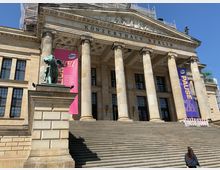 Das Bild zeigt die Fassade des Konzerthauses Berlin mit symmetrischen Säulen im klassizistischen Stil. Vor dem Gebäude befindet sich eine breite Treppe, und rechts und links hängen bunte Banner; links in Pink und rechts in Lila. Im Vordergrund steht eine grüne Skulptur auf einem Podest.