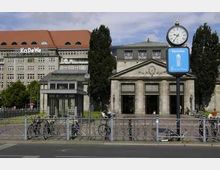 Historische Eingangshalle mit Säulen neben einem modernen Geschäftshaus versehen mit der Aufschrift „KaDeWe“. Im Vordergrund stehen Fahrräder an einem Geländer, und eine Straßen-Uhr ist sichtbar.