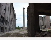 Industriegelände mit verlassenen, teilweise beschädigten Gebäuden. In der Mitte ragt ein hoher Schornstein auf, umgeben von Trümmern und überwucherten Flächen.