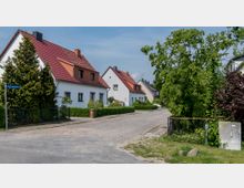 Straßenszene in einem Wohngebiet mit Einfamilienhäusern, die weiße Fassaden und rote Ziegeldächer haben. Die Straße ist gepflastert, von Bäumen und Büschen umgeben, und ein Straßenschild weist auf die „Friedrich-Ludwig-Jahn-Straße“ hin.