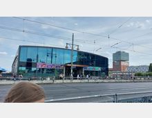 Außenansicht des Bahnhofs Warschauer Straße in Berlin, einem modernen Gebäude mit großen Glasfassaden. Im Vordergrund sind Straßenbahnschienen und eine Fußgängerbrücke, während im Hintergrund weitere Gebäude, darunter ein Hochhaus, zu sehen sind.