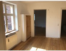 Leerer Raum mit Holzboden, weiß gestrichenen Wänden und zwei Türen, die zu weiteren Räumen führen. Links befindet sich ein großes, weiß gerahmtes Fenster mit Ausblick auf eine Ziegelwand, sowie eine Heizung darunter.