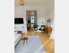 Wohnzimmer mit Holzboden, modern eingerichtet mit einem weißen Sofa, einem kleinen runden Beistelltisch und einem Zickzack-Teppich. Im Hintergrund ist ein Essbereich mit Hängelampen sichtbar, der durch einen breiten Durchgang mit weißen Vorhängen abgetrennt ist.