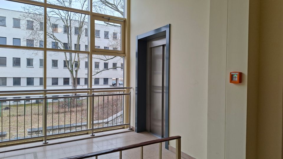 Innenflur eines modernen Gebäudes mit Aufzugtür in einem dunklen Rahmen und großflächigen Fensterfronten. Vor den Fenstern verläuft ein Metallgeländer; draußen sind ein Innenhof mit Bäumen und ein gegenüberliegendes mehrstöckiges Gebäude zu sehen.