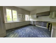 Geräumige Küche mit weißen Einbauküchenmöbeln, großen Fenstern, blauem Fliesenboden und weißen Wandfliesen. Auf der rechten Seite befindet sich ein großer Edelstahlherd mit Abzugshaube.