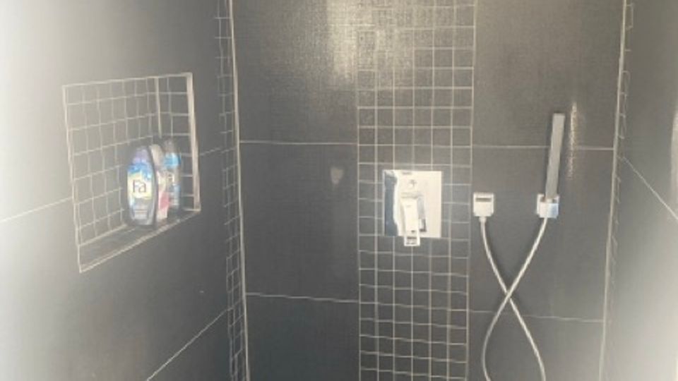 Geflieste, bodengleiche Dusche mit dunklen Wandfliesen und einem vertikalen Streifen aus kleinen Mosaikfliesen in der Mitte. Sichtbar sind eine Regendusche an der Decke, eine Handbrause mit Schlauch sowie eine eingelassene Wandnische mit Ablage.