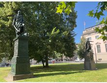 Zwei Denkmäler stehen auf einer Grünfläche mit großen Bäumen im Hintergrund. Ein klassisches Gebäude mit heller Fassade und geometrischen Fenstern sowie eine deutsche Flagge auf dem Dach sind im Hintergrund sichtbar.