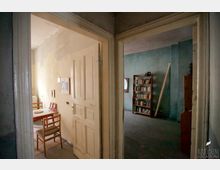 Blick in zwei Räume eines Altbaus mit abgenutzten Wänden: Links ein beige gestrichener Raum mit Holztisch und Stühlen, rechts ein blau gestrichener Raum mit einem Regal voller Bücher und einem runden Wandspiegel.