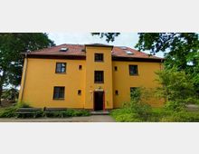 Gelbes Gebäude mit drei Etagen, rotem Satteldach und zentralem Eingang in einem kleinen Vorsprung. Vor dem Gebäude stehen Bänke, umgeben von Bäumen und Sträuchern in einer parkähnlichen Umgebung.