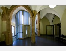 Innenansicht eines historischen Gebäudes mit spitzbogigen Steinsäulen, gotischen Gewölben und modernen Elementen wie einer verglasten Aufzugskabine. Im Hintergrund sind verzierte Fenster mit Gittermustern sowie eine Glastür erkennbar.