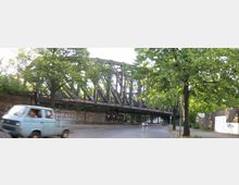 Eine alte Eisenbahnbrücke mit Stahlträgern, teilweise mit Graffiti bedeckt, überspannt eine gepflasterte Straße, die von Bäumen gesäumt ist. Im Vordergrund fährt ein blauer Transporter, während im Hintergrund ein Auto unter der Brücke hindurchfährt.