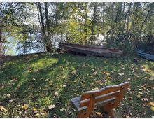 Eine hölzerne Bank steht auf einer Wiese, umgeben von herbstlichem Laub und Bäumen. Im Hintergrund ist ein altes Holzboot neben dichtem Gebüsch am Ufer eines Sees zu sehen.
