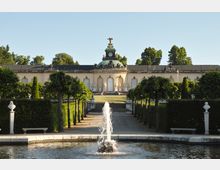 Barockes Gebäude mit zentralem Uhrturm und kupfergrünem Dach, umgeben von einer symmetrischen Gartenanlage mit geschnittenen Bäumen und Skulpturen. Im Vordergrund ein Wasserbecken mit einer sprudelnden Fontäne.