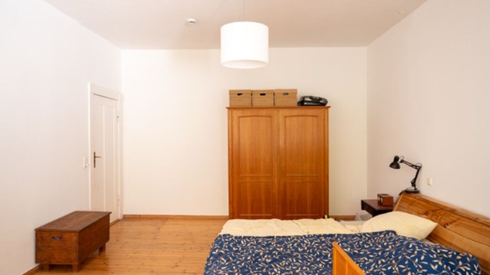 Schlichtes Schlafzimmer mit Holzboden, Doppelbett mit blau gemusterter Tagesdecke und Holz-Kopfteil sowie einer Deckenleuchte in der Mitte. An der Wand stehen ein großer Holzkleiderschrank und eine Truhe, daneben eine weiße Zimmertür und eine Wandlampe am Bett.