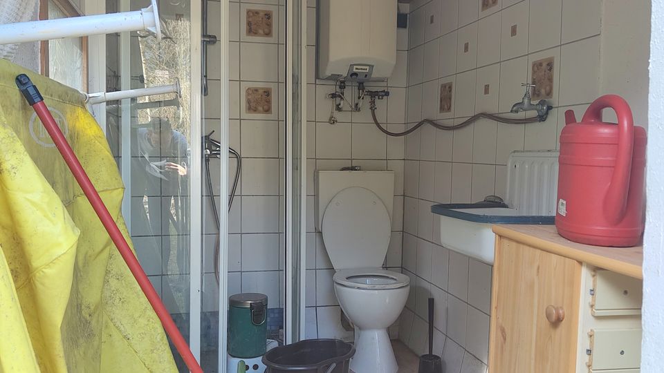 Schmaler, gefliester Sanitärraum mit Holzdecke, Duschkabine links und Toilette am Ende; an der rechten Wand stehen ein kleines Waschbecken mit Unterschrank, ein Heizkörper und ein Wandgerät. Im Vordergrund sind Putzutensilien wie Eimer, Mop und Gießkanne sowie eine rote Thermoskanne auf dem Schrank zu sehen.