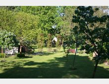 Grüne Gartenlandschaft mit gepflegtem Rasen, blühenden Bäumen und Sträuchern. Im Hintergrund sind metallene Rankgerüste mit Pflanzen sowie ein kleines, teilweise sichtbares Gebäude mit weißer Fassade zu sehen.