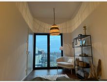 Gemütlich eingerichtetes Zimmer mit einem großen Fenster und Zugang zu einem Balkon, im Hintergrund ist eine städtische Aussicht zu sehen. Der Raum ist mit einer hellen Couch, einem schwarzen Regal, einem Hängelampenschirm aus Rattan und Trockenblumen dekoriert.