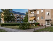 Mehrfamilienhäuser in einer Wohnsiedlung mit einfachen Fassaden, Balkonen und Blumenbepflanzung, flankiert von gepflasterten Wegen, niedrigem Gebüsch und Grünflächen. Eines der Häuser hat Solarzellen auf dem Dach.