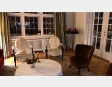 Gemütliches Wohnzimmer mit Holzboden, einer Sitzgruppe aus weißen und braunen Polsterstühlen sowie einem runden Tisch mit weißem Tischtuch. Im Hintergrund sind eine große Fensterfront mit Sprossenglas, grüne Vorhänge, Pflanzen und ein dekorierter Adventskranz zu sehen.