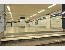 Unterirdischer Durchgang mit hellen Wänden aus weißen und blauen Fliesen, rechteckigen Säulen und intensiver Deckenbeleuchtung. Im Vordergrund führen Stufen hinab, im Hintergrund sind Türen und Werbeplakate sichtbar.