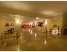 Geräumiger Eingangsbereich eines Gebäudes mit gefliesten Böden, hellbeigen Wänden und Wandlampen. Rechts ist eine Treppe sichtbar, links ein Handlauf, ein kleiner Tisch mit Stühlen, und im Hintergrund leuchtet eine rote, dekorative Wandverkleidung.