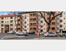 Wohngebäude aus der Nachkriegszeit mit hellbeigem Putz und rotbraunen Backsteinelementen, mehreren Balkonen und großen Fenstern, gelegen an einer Straßenecke mit geparkten Autos und einem Stoppschild. Im Vordergrund stehen kahle Bäume und eine orangefarbene Mülltonne.