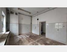 Ehemaliger leerer Raum mit abgenutztem Boden und gefliesten Wänden, teilweise beschädigt. Die Decke zeigt Abnutzungsspuren, und ein Türrahmen führt in einen angrenzenden Bereich.