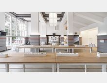 Großer, heller Innenraum mit hohen Decken und modernen, weißen Wänden, unterbrochen von grauen Pfeilern mit roten Streifen. Im Vordergrund stehen Holztische, während im Hintergrund kleinere Tischgruppen mit weißen Tischdecken vor großen, bodentiefen Fenstern platziert sind.