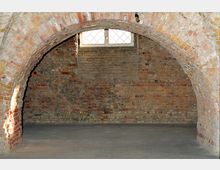 Kellergewölbe aus roten und beigen Ziegelsteinen mit einem Rundbogen aus Mauerwerk im Vordergrund; dahinter eine kleine Fensternische mit einem vergitterten Fenster, das Tageslicht hereinlässt, sowie ein Betonboden.
