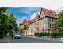 Straßenszene mit einem großen, historisch wirkenden Gebäude mit gelblich-beigem Putz und rotem Ziegeldach, das von mehreren Giebeln und einem Turm geprägt ist. Im Vordergrund sind ein Gehweg, parkende Autos und Fahrräder zu sehen, umgeben von Bäumen und grüner Vegetation.