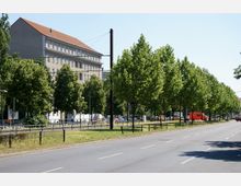 Straßenansicht mit einer breiten Fahrbahn, gesäumt von einer Baumreihe und einzelnen Laternen. Im Hintergrund steht ein mehrstöckiges Gebäude mit rotem Dach, während am Straßenrand parkende Autos und ein orangefarbener Transporter zu sehen sind.