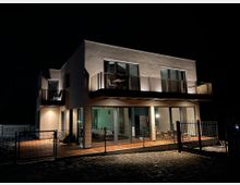 Ein modernes zweistöckiges Wohnhaus bei Nacht, mit großen Fensterfronten und Balkon im oberen Stockwerk. Der Eingangsbereich und die Terrasse sind durch Außenbeleuchtung erhellt, umgeben von einem Metallzaun auf einem gepflasterten Boden.