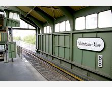 Bahnhof Schönhauser Allee mit grün gestrichenen Stahlwänden und großen Fenstern, Blick auf die Gleise und das Plattformdach. Ein Schild mit dem Namen "Schönhauser Allee" ist an der Wand befestigt.