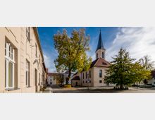Historisches Stadtbild mit einer kleinen Kirche mit spitzem Turm und roten Dachziegeln, umgeben von Bäumen im Herbstlaub. Links im Bild eine Reihe beige verputzter Gebäude mit weißen Fensterrahmen, rechts ein gepflasterter Platz.