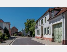 Straßenszene in einem kleinen Dorf mit Kopfsteinpflaster, historisch anmutenden Gebäuden mit roten Ziegeldächern und pastellfarbenen Fassaden. Rechts im Bild ein Haus mit grünen Fensterläden und einem großen Tor, links eine Baumreihe und Blumenbeete.